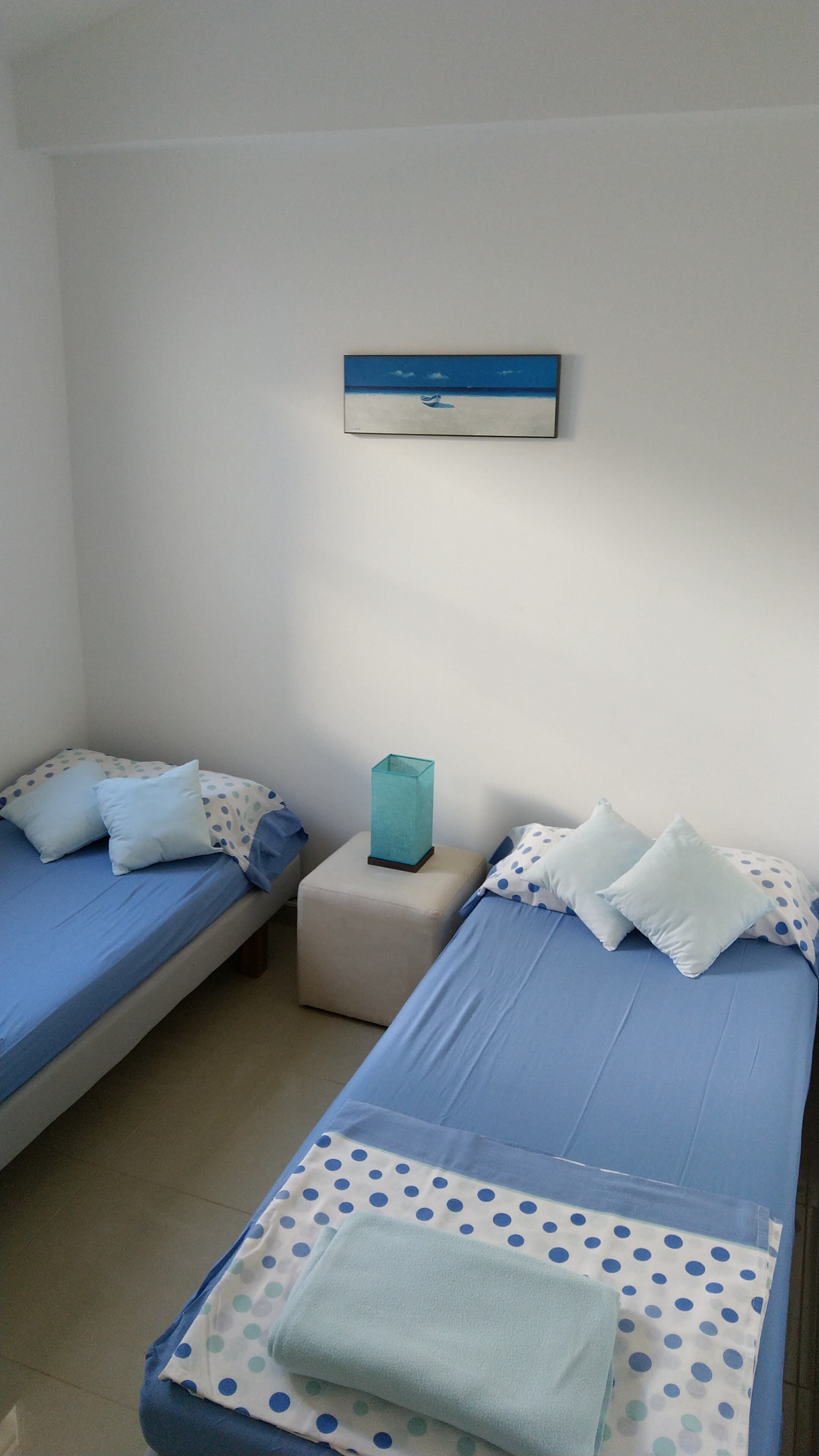 A room with two single beds, each with blue bedding and white pillows with blue polka dots. There is a small table with a teal vase and a white pillow. A framed picture hangs on the wall above the beds.