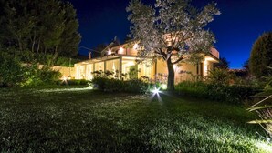 Une maison bien éclairée, avec un grand arbre dans le jardin, entourée d’une végétation luxuriante.