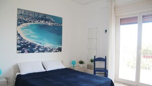 Ein Schlafzimmer mit Bett, Nachttisch, Sessel, Fenster und einem Bild einer Küstenlandschaft.