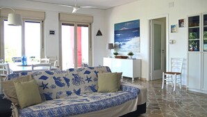 Ein Wohnzimmer mit einem gemusterten Sofa, einem Esstisch, Stühlen, einem Deckenventilator, einem Gemälde und einem Bücherregal.