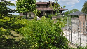 Une maison à deux étages, avec un toit en tuiles, entourée d’une végétation luxuriante et d’une clôture en métal.