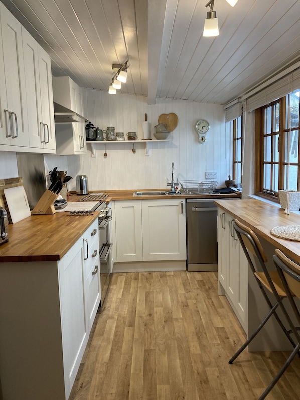 A kitchen with white cabinets, wooden countertops, and a stainless steel dishwasher. There are shelves with jars and a clock on the wall.