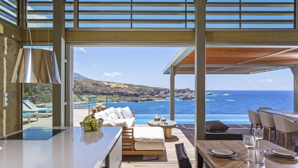 Une cuisine moderne avec vue sur l’océan, un coin repas avec une table dressée pour deux personnes, et un espace salon confortable avec un canapé et des coussins.