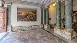 Un vaste hall d’entrée avec un sol en marbre, des colonnes de style classique, un grand tableau représentant un paysage et un meuble en bois.
