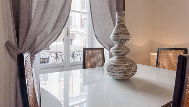Une table à manger avec un vase, des rideaux et une vue sur des immeubles par la fenêtre.