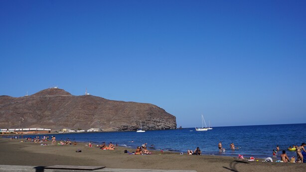 A beach with people sunbathing and swimming, a sailboat, and a rocky hill in the background. - Apartment next to the beach with free Wifi, SAT TV, sea views (Gran Tarajal - Fuerteventura)