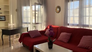A living room with a red sofa, a white coffee table, and a small table with a plant. There is a window with sheer curtains and a view of the outside. - Apartment next to the beach with free Wifi, SAT TV, sea views (Gran Tarajal - Fuerteventura)