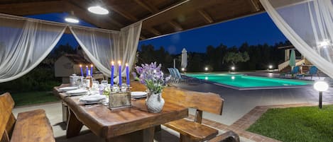 An outdoor dining area with a wooden table set for a meal, surrounded by chairs, and a pool in the background.