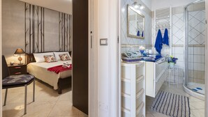 Une chambre à coucher comprenant un lit, une table de chevet, une lampe et une chaise. À côté se trouve une salle de bain avec une douche, un lavabo, un miroir et un porte-serviettes.