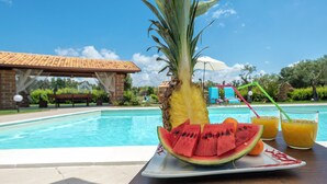 Une table placée au bord de la piscine, avec un ananas, des tranches de pastèque et deux verres de jus d’orange.