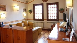A cozy living room with a wooden coffee table, a sofa, and a TV. There are two windows with decorative glass panes and wall-mounted lamps.