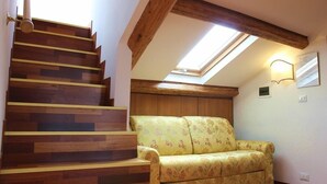 A wooden staircase leading to a loft bedroom with a skylight, a floral sofa, and a wall-mounted light fixture.