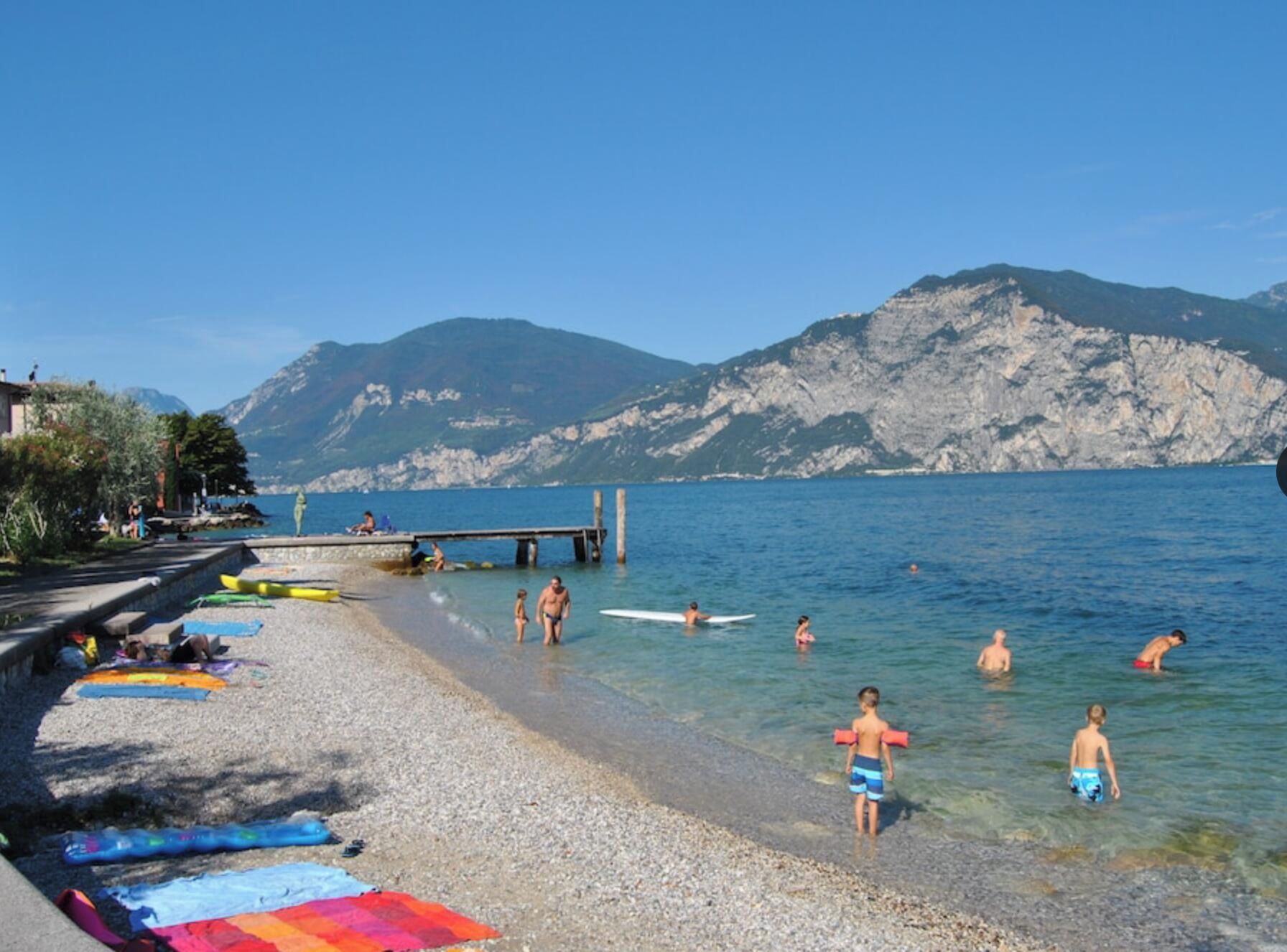 Une plage de galets bordée d’une eau cristalline, avec une jetée et des montagnes en arrière-plan.