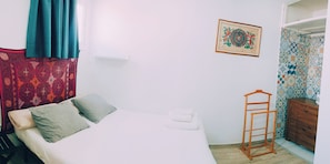 Un lit bien fait, agrémenté d’oreillers, un porte-vêtements suspendu et une petite commode en bois dans une pièce dont le mur est décoré d’un motif et qui possède une porte.