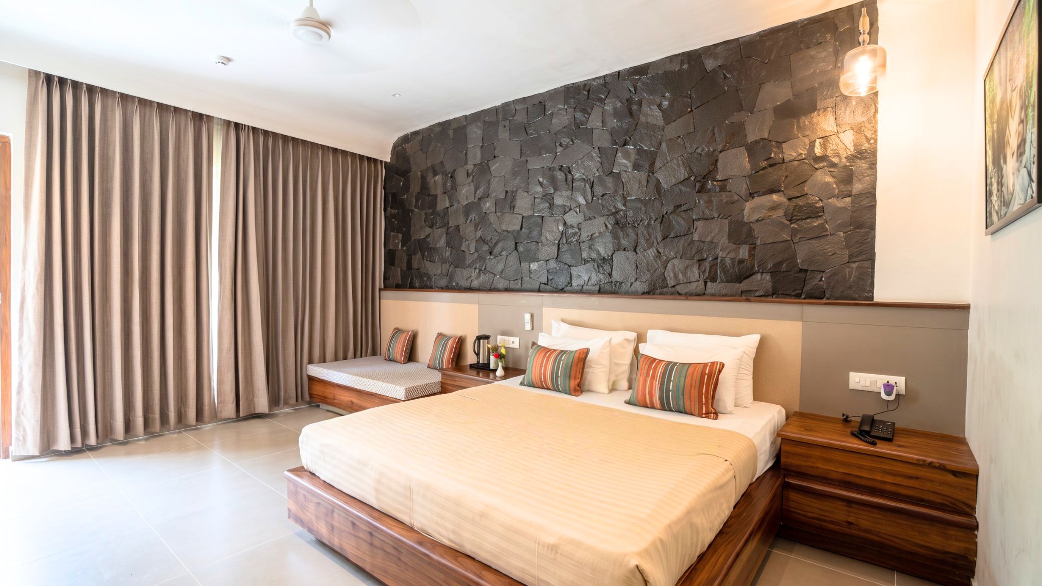 Una habitación de hotel moderna con una cama grande, una pared de acento de piedra, una mesita de noche con teléfono y una ventana con cortinas.
