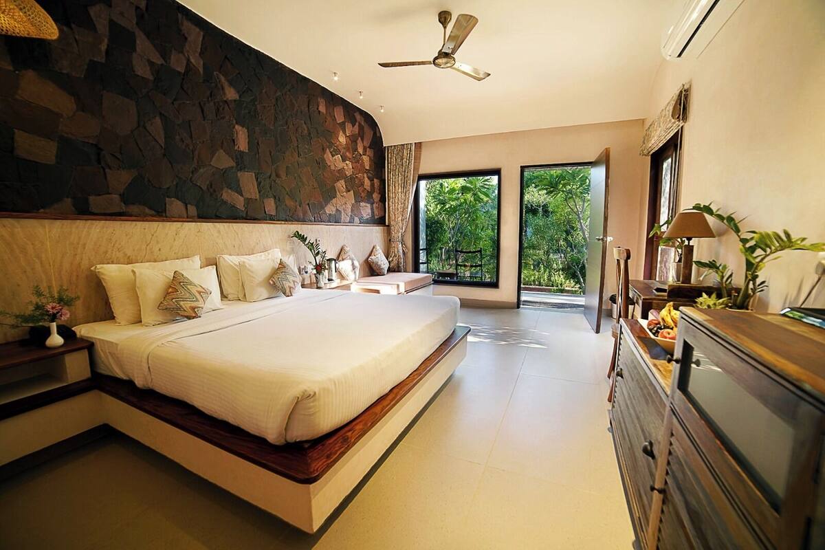 Une chambre moderne avec un grand lit, un mur décoratif en pierre, un ventilateur de plafond et une vue sur la verdure par la fenêtre.