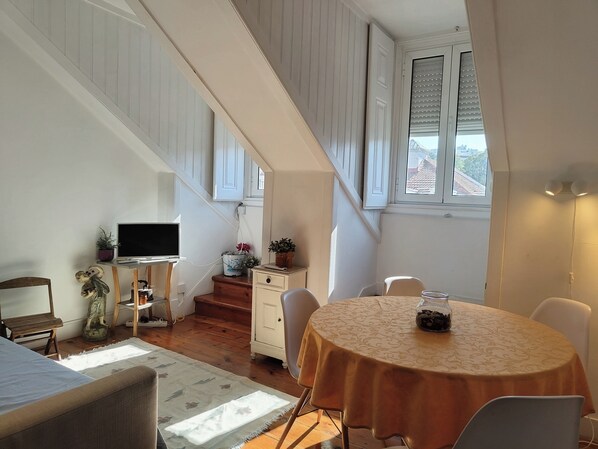 A room with a round table, a TV, a staircase, and a window with blinds.