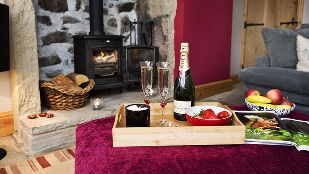A cozy living room with a stone fireplace, a wooden table with a bottle of champagne, glasses, strawberries, and a bowl of fruit. - Relax in the Heart of Bronte Country (Haworth)