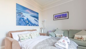 A hotel room with a bed, a green sofa, a large painting of a mountain landscape, and a smaller framed picture of a cityscape.