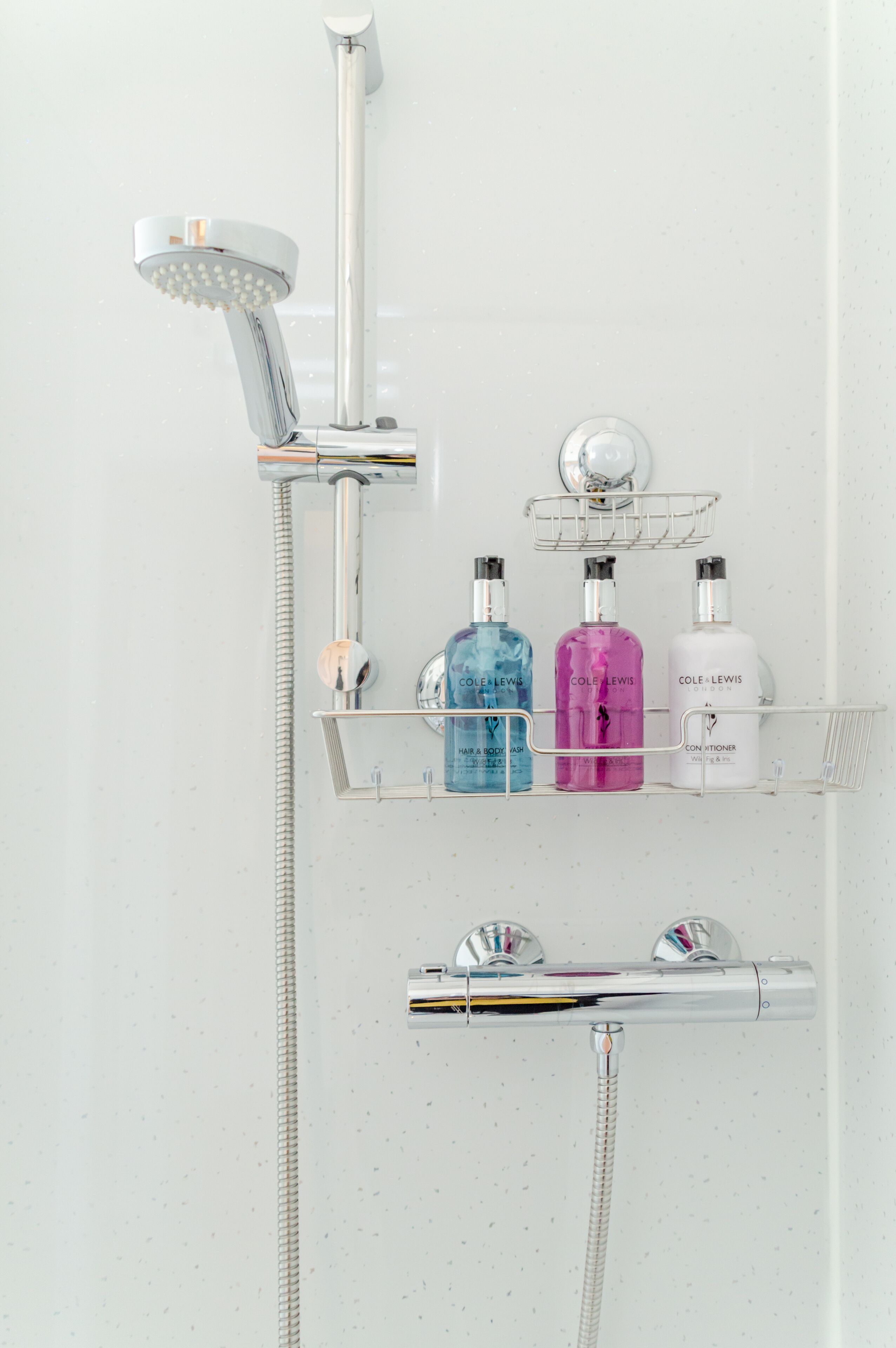 Une douche équipée d’un pommeau de douche à main et d’un robinet mural. Trois flacons de produits de toilette sont posés sur une étagère.