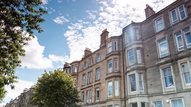 A row of traditional terraced houses with uniform windows and doors, a clear sky, and a tree in the foreground.