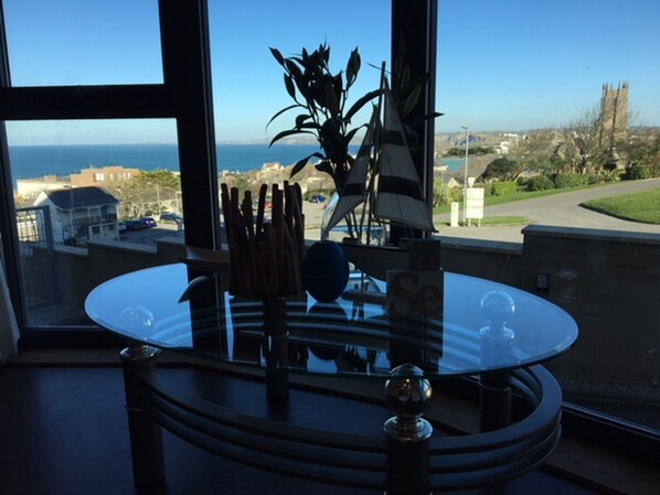 A dining area with a glass table, a plant, and a view of the sea and buildings.