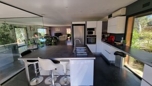 Een moderne keuken met een centraal eiland, barstoelen, een inbouwoven en een groot raam met uitzicht op groen.