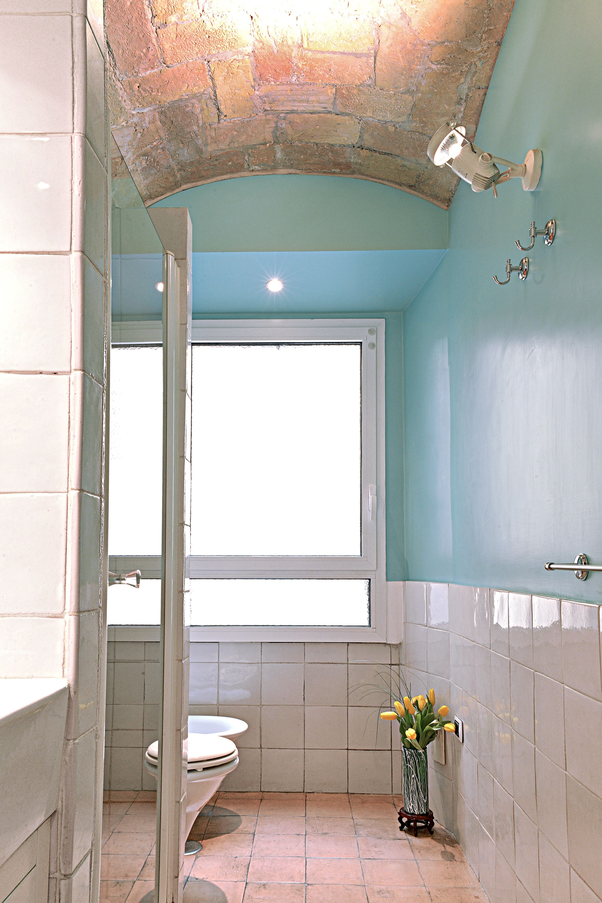 A bathroom with a stone arch ceiling, a window, a toilet, and a vase with flowers.