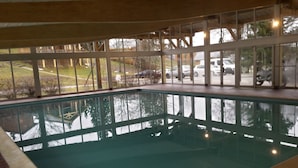 Una piscina interior con paredes de vidrio transparente, borde de ladrillo y vistas a coches aparcados y árboles en el exterior.