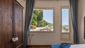 Un dormitorio con un armario de madera, una cama con una manta azul, un ventanal con vistas a muebles de exterior y follaje, y un soporte metálico con tela blanca.
