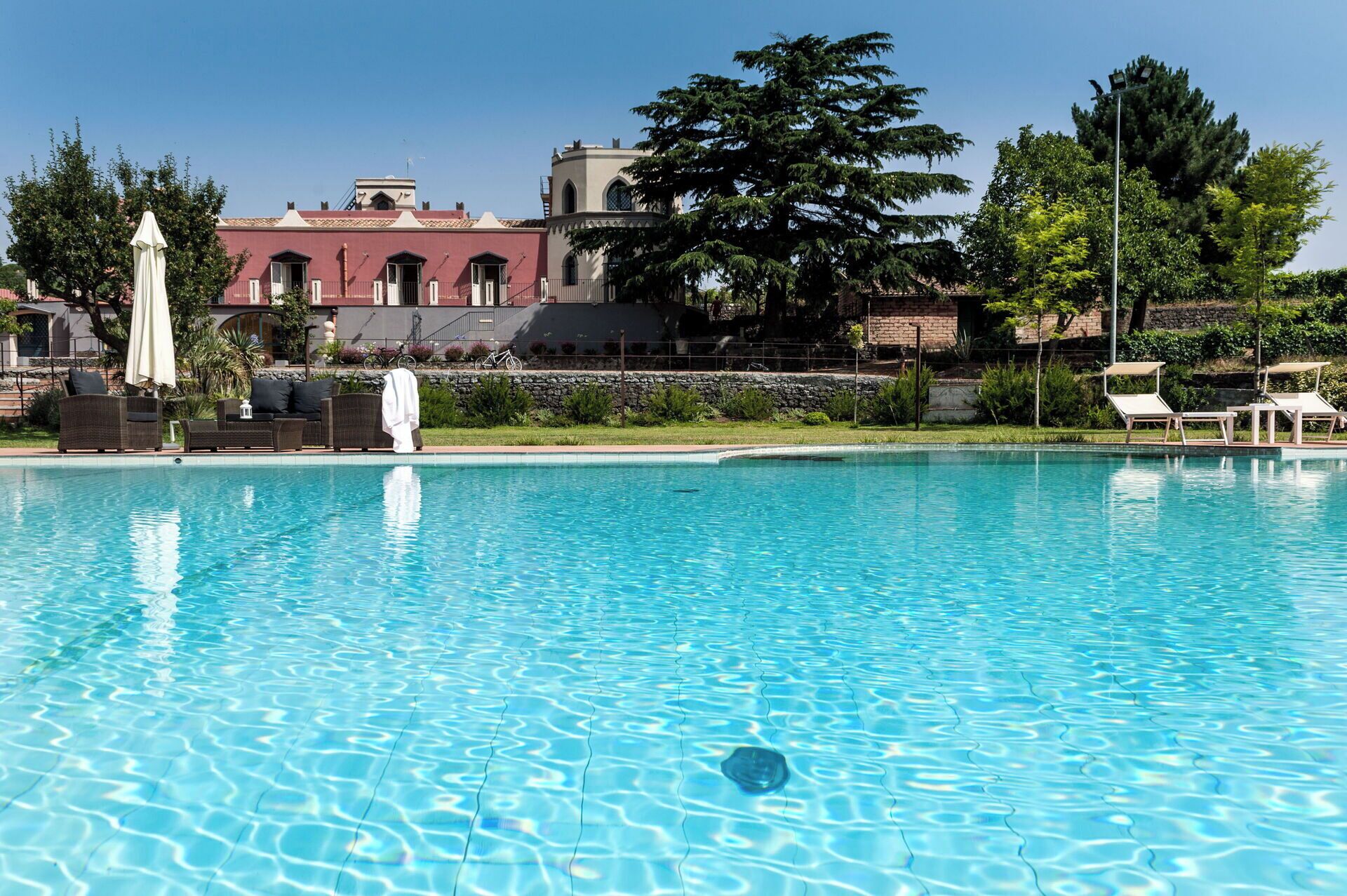 A swimming pool with lounge chairs and umbrellas, surrounded by greenery and a building with a red facade.