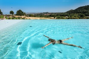 Une piscine d’eau cristalline où une personne nage, entourée d’une plage de sable et de verdure.