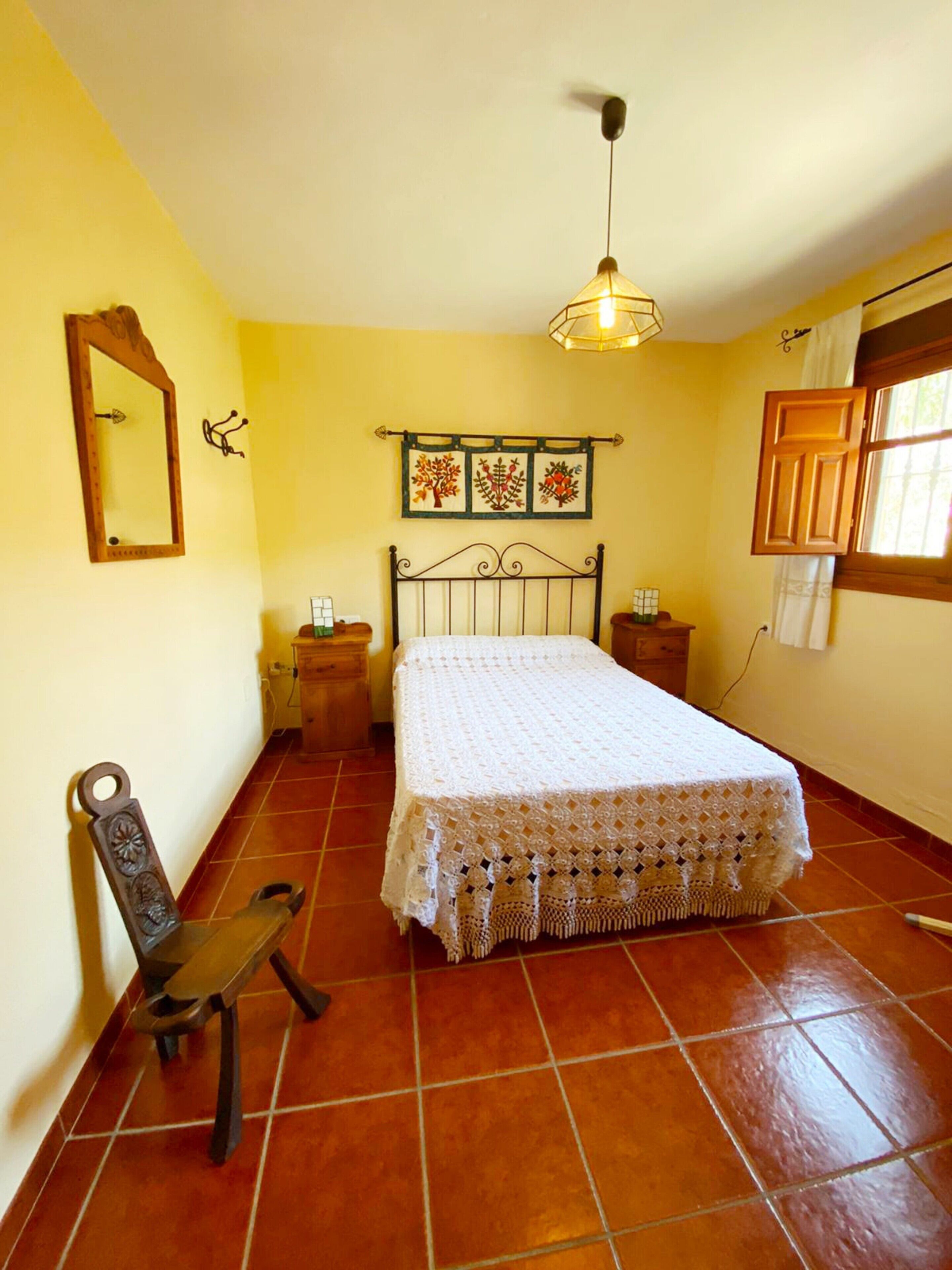 A bedroom with a bed, a chair, a nightstand, a hanging light fixture, a mirror, and a window with curtains.