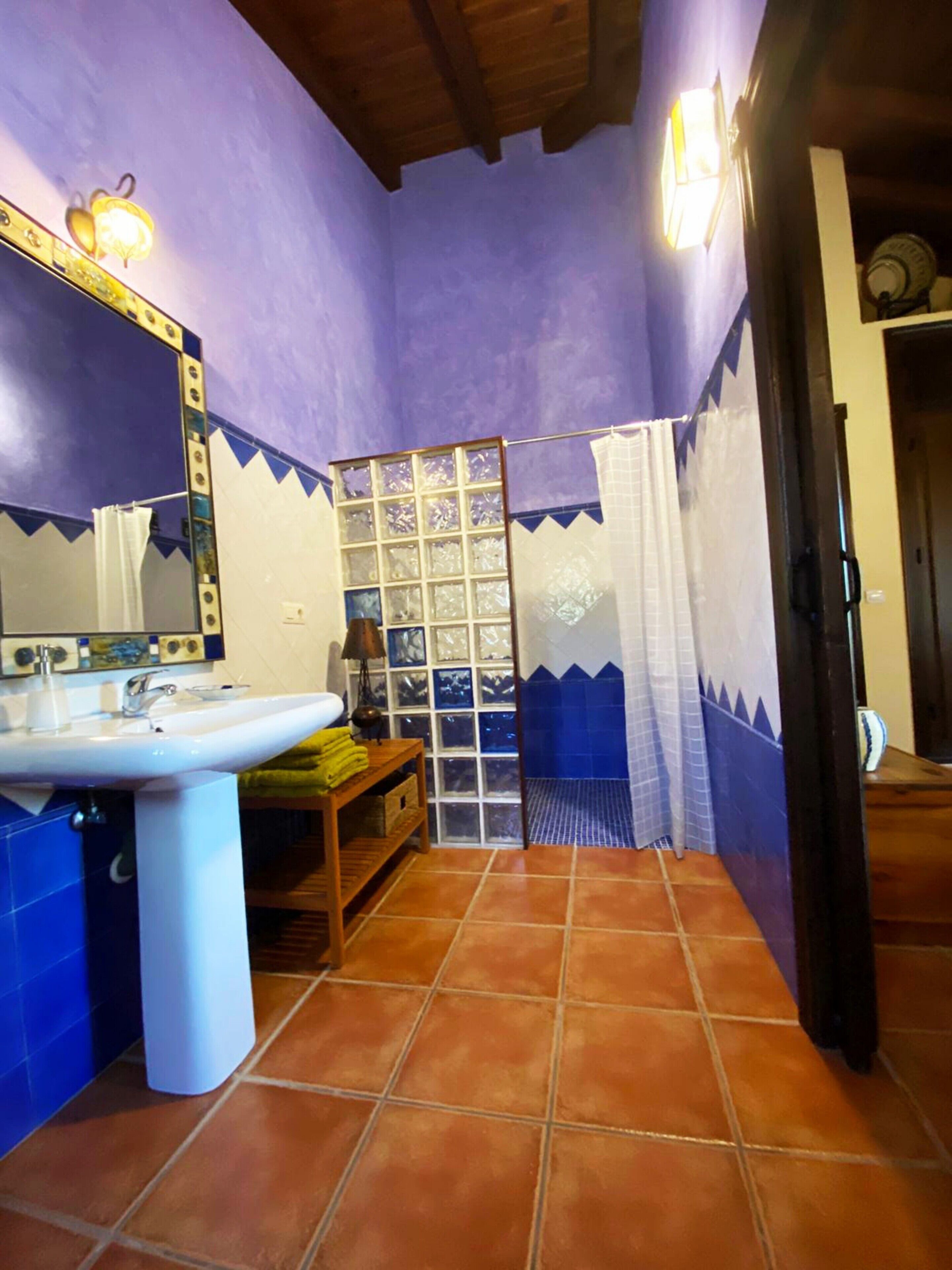 A bathroom with a white sink, a mirror with lights, a window with a grid pattern, and a shower area with blue and white tiles.