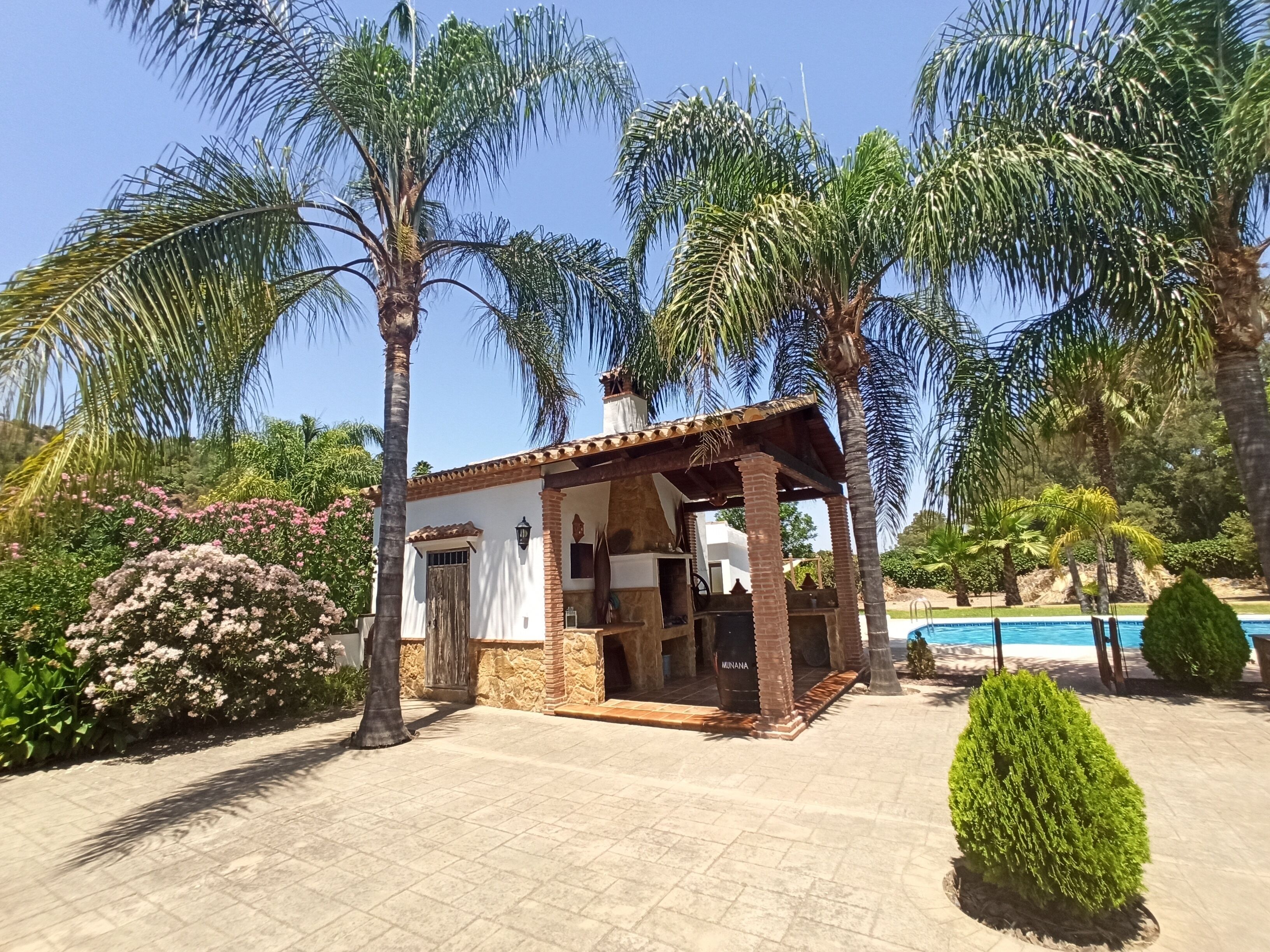 A small, rustic building with a tiled patio, surrounded by palm trees and a pool.