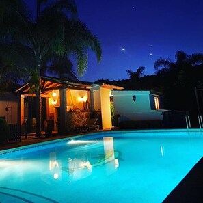 A nighttime pool area with a lounge chair and a small building with lights.