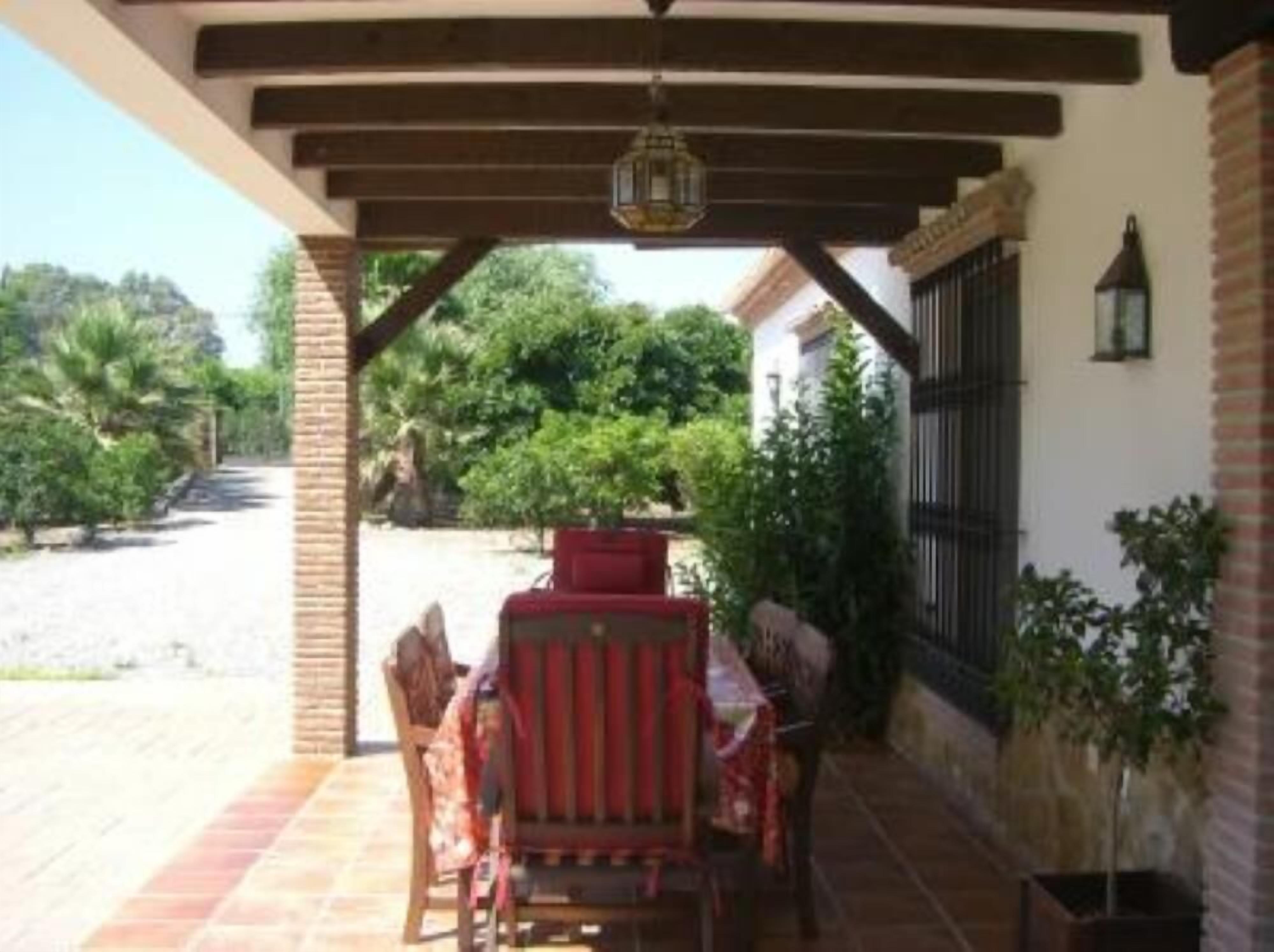 An outdoor patio area with a table and chairs, a hanging lantern, and a view of a garden with trees and a building in the background.