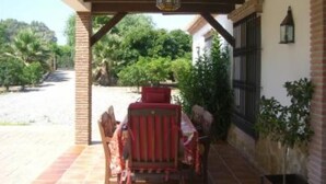 An outdoor patio area with a table and chairs, a hanging lantern, and a view of a garden with trees and a building in the background.