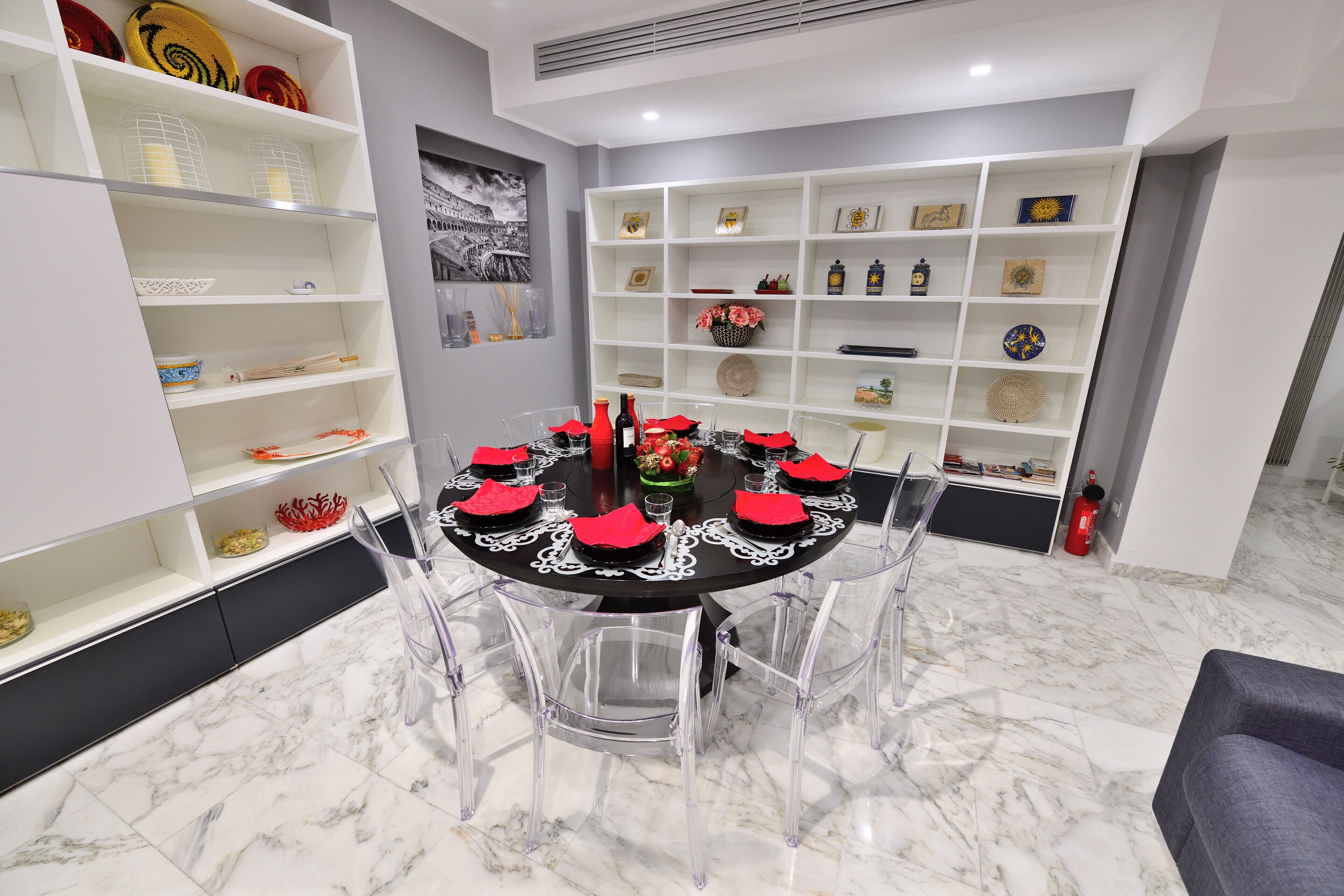 Un coin repas avec une table ronde dressée pour six personnes, entourée de chaises transparentes. Des étagères ornées d’objets décoratifs bordent les murs.