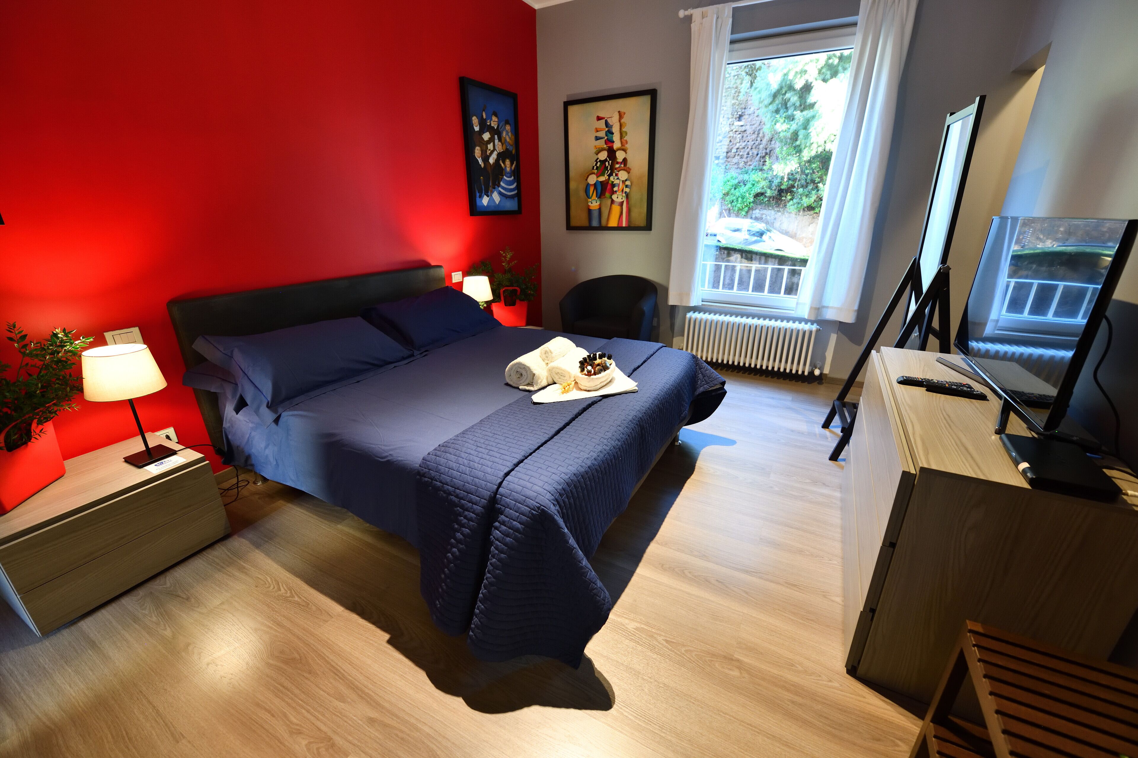 A hotel room with a bed, a desk, a chair, a lamp, and a window with a view of greenery.