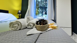 Un lit bien fait, avec une serviette blanche roulée, un panier contenant des articles de toilette et un bol de grains de blé, sur une table de chevet dans une chambre d’hôtel.