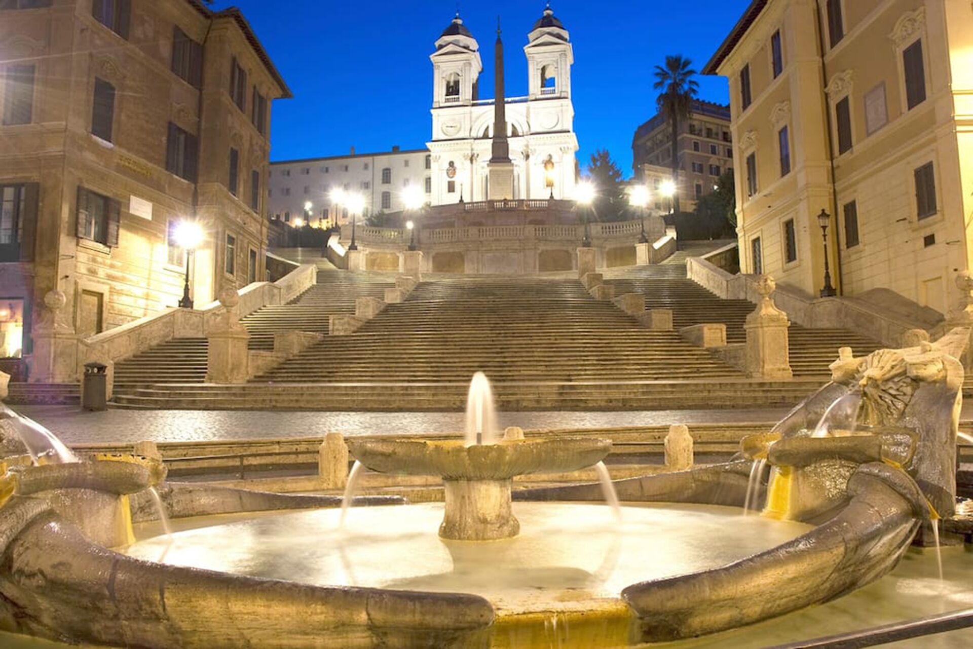 Un grand escalier mène à une église blanche dotée de deux tours, qui est entourée de bâtiments historiques et d’une fontaine centrale.