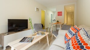 A modern living room with a flat-screen TV, a coffee table with magazines, a sofa with patterned cushions, and a dining area with chairs and a table. - Brand New Apartment Near Camp Nou & Fira Barcelona (L'Hospitalet de Llobregat)