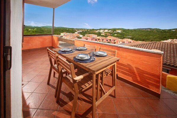 Mesa de madera dispuesta con platos y vasos en un balcón con baldosas de terracota, con vista a una zona residencial con casas y colinas verdes.
