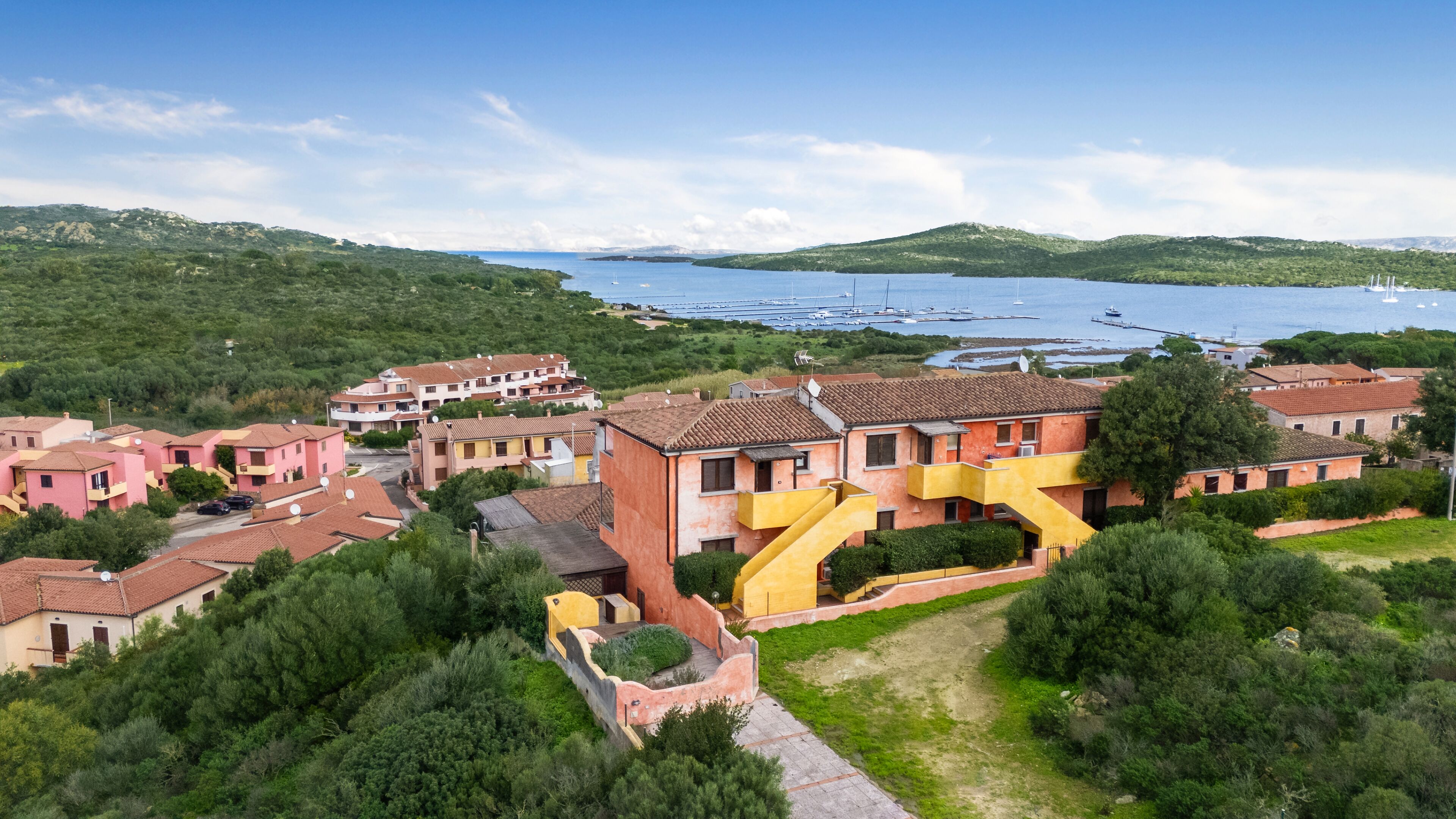 Uma vila costeira com casas em terraços, um edifício amarelo destacado e uma marina com barcos.