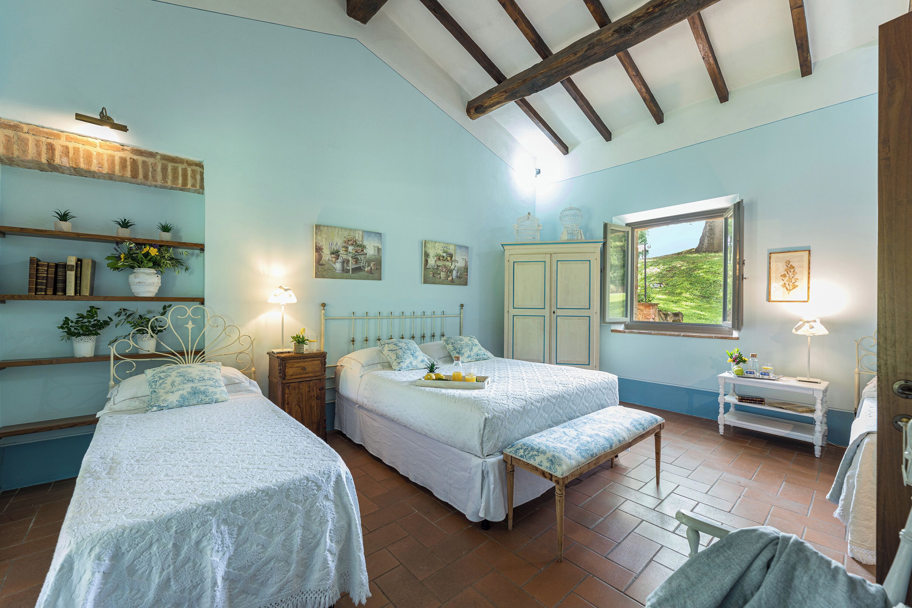 A bedroom with two beds, a wooden bench, a bedside table with a lamp, a shelf with books and plants, and a window with a view of greenery.