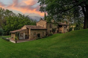 Une maison en pierre avec un toit de tuiles, entourée d’une végétation luxuriante et d’arbres.
