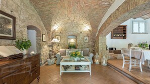 A cozy room with a stone wall, a wooden dresser, a glass coffee table, a sofa, and a dining area with a table and chairs. - Il Molinaccio 14+2, Emma Villas (Montepulciano (Siena))
