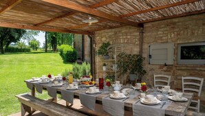 An outdoor dining area with a long wooden table set for a meal, surrounded by brick walls and a thatched roof. - Il Molinaccio 14+2, Emma Villas (Montepulciano (Siena))