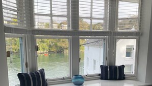 Une fenêtre en saillie avec des stores blancs, un vase bleu et deux coussins à rayures.